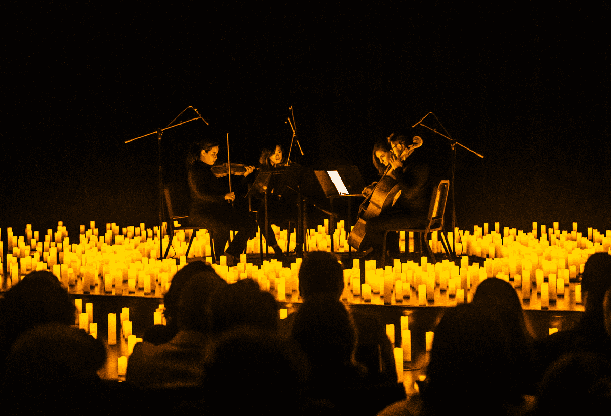 Behind the silhouette of an audience a string quartet is performing on a raised stage covered in candles.