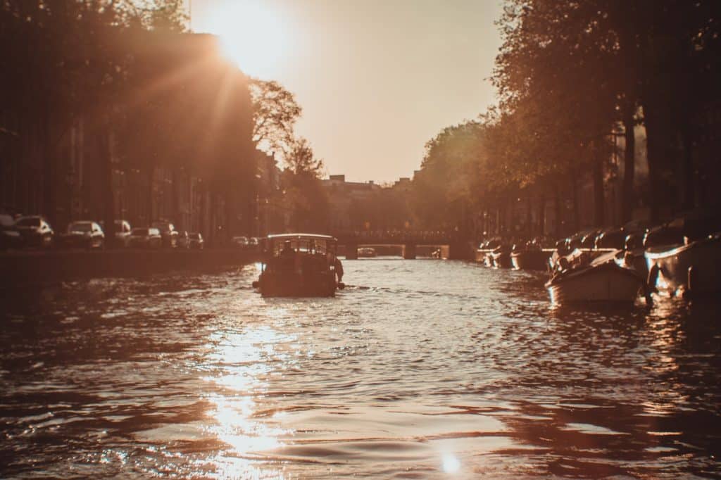 A boat sails down a canal in Amsterdam in warm weather, with the dun shining behind it.