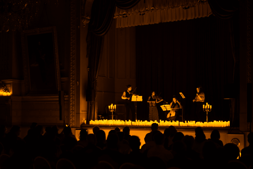 The silhouette of an audience watching a string quartet perform on a raised stage covered in candles.