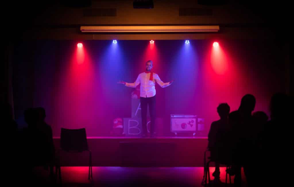 A comedian performs on a stage lit with red and blue spotlights.